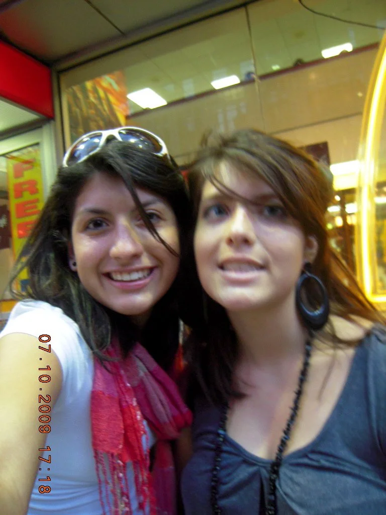 Two young women taking a selfie in front of a store window, smiling at the camera.