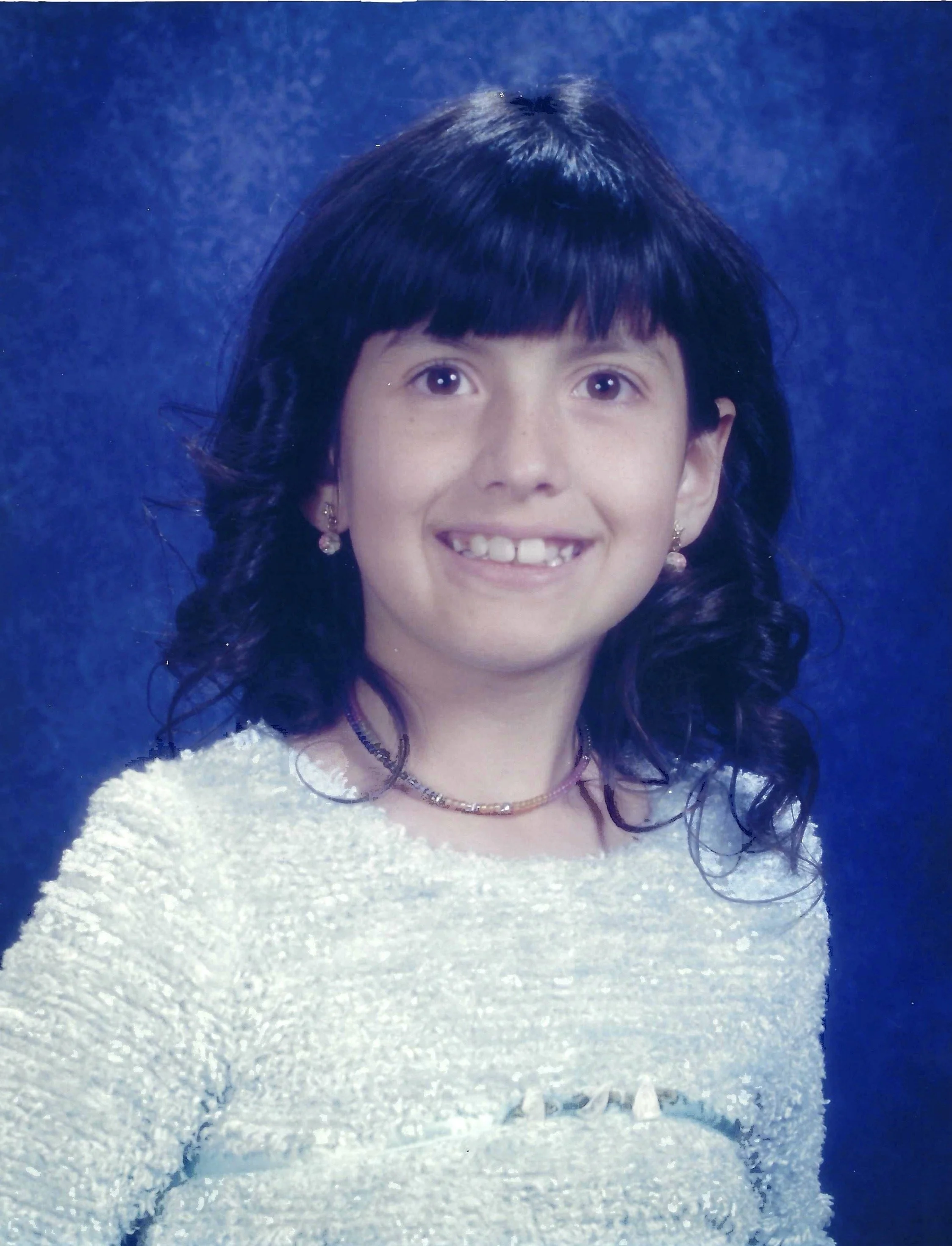 A young girl with dark hair, blunt bangs, and a big smile, standing in a snowy landscape with a blue background.