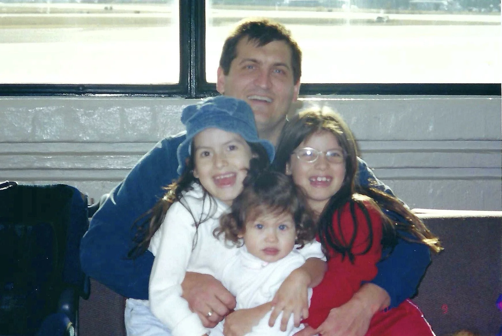 A family of five sitting on a bench at an airport, smiling at the camera. The man is in the back with three young girls in front of him, all wearing bright clothing and showing happy expressions.