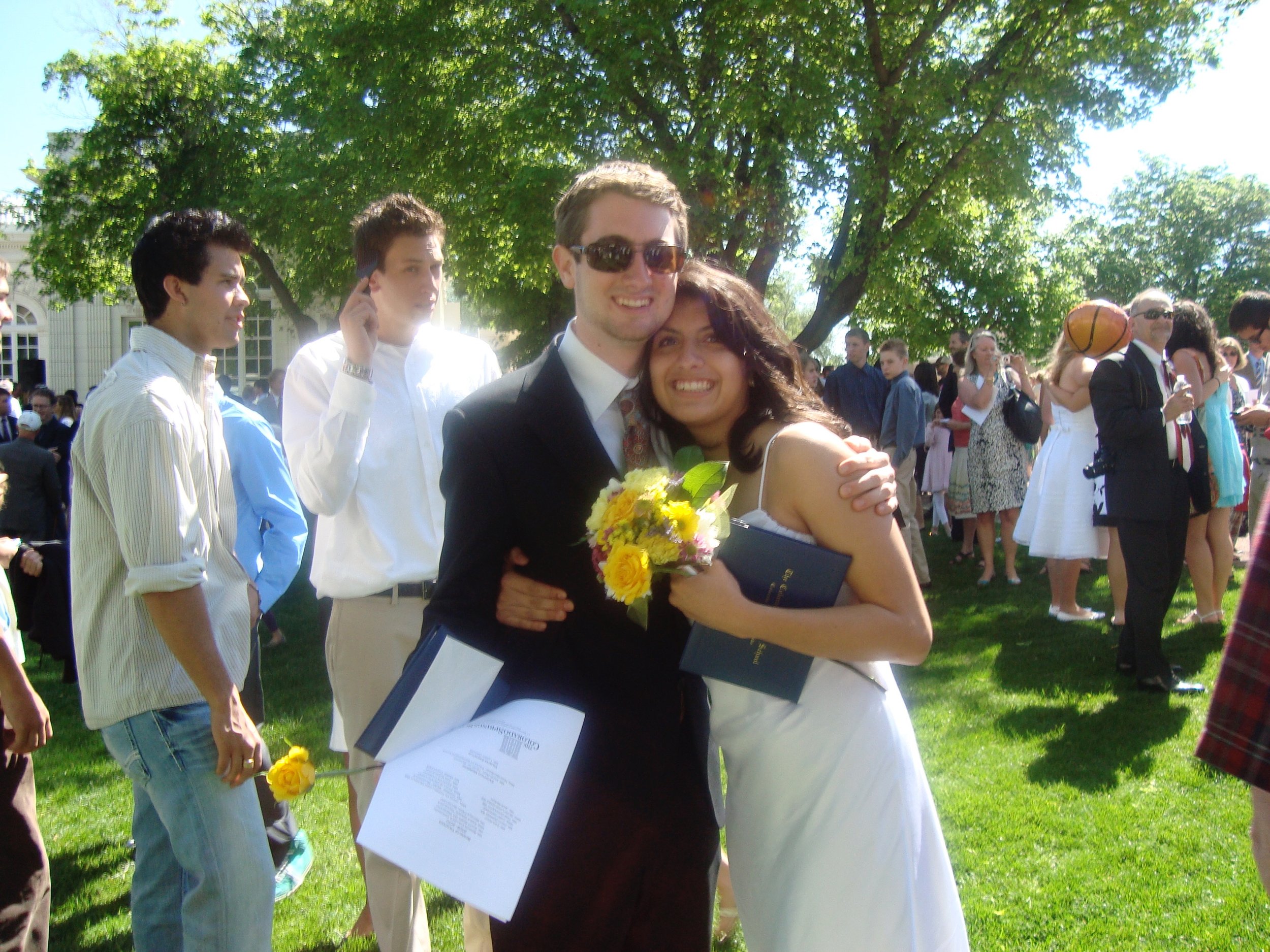 A joyful young couple hugging at an outdoor event, with the man wearing sunglasses and holding a bouquet of yellow flowers, surrounded by other people in formal and casual attire, under green trees on a sunny day.