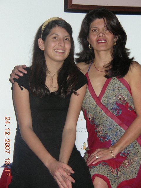 Two women standing side by side indoors, smiling at the camera. The woman on the left has long dark hair, is wearing a black dress, and a beige headband. The woman on the right has dark hair with bangs, is wearing a colorful dress with red, purple, a