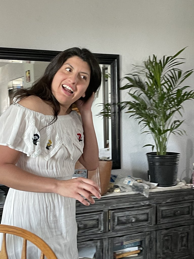 A woman with dark hair, wearing a white dress with embroidered bird designs, stands indoors and looks surprised or amused while holding a glass of drink, with a large green potted plant and a black-framed mirror behind her.
