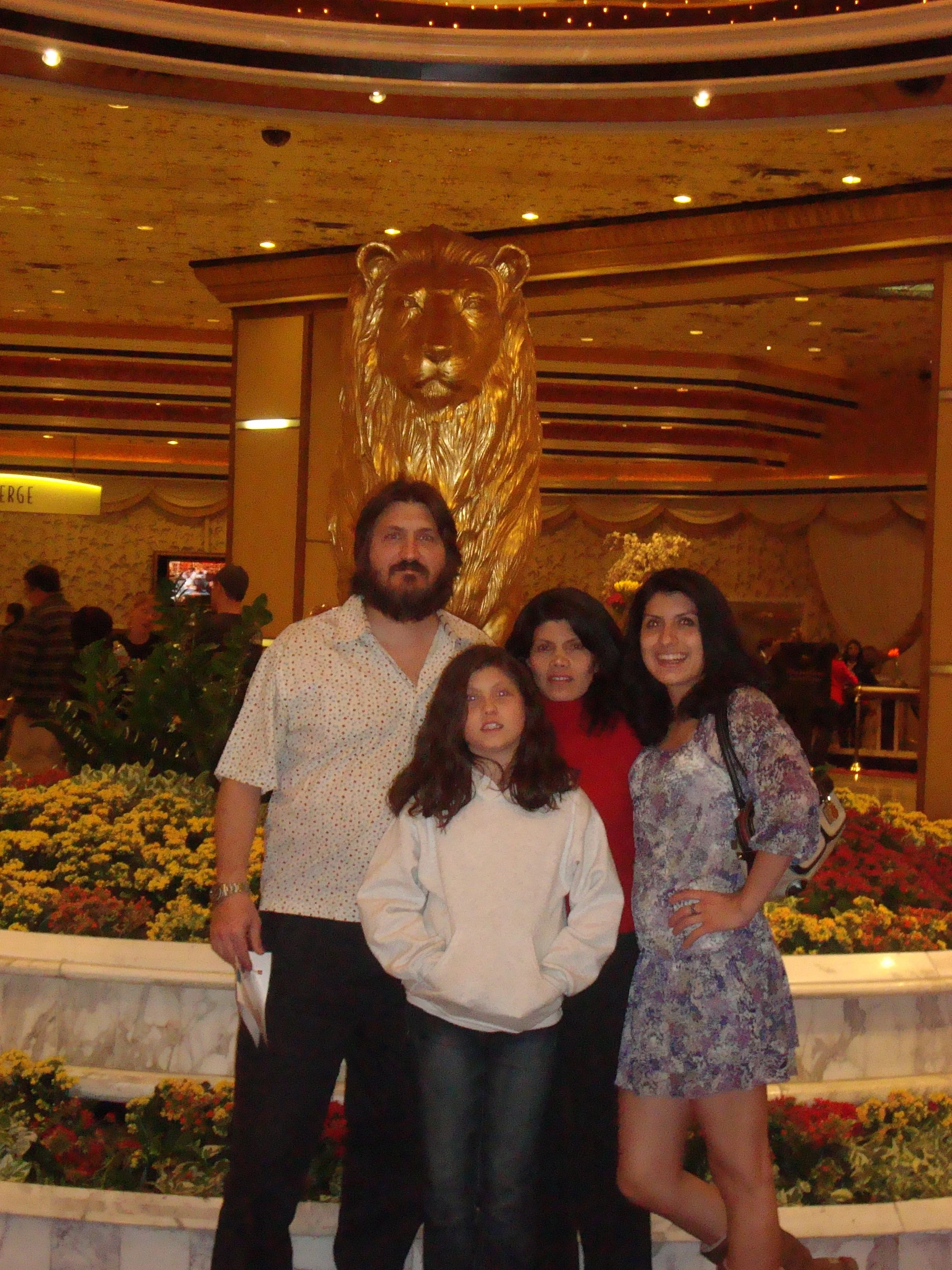 Group of five people posing in front of a large golden lion statue inside a building. The background shows a decorated ceiling and floral arrangements.