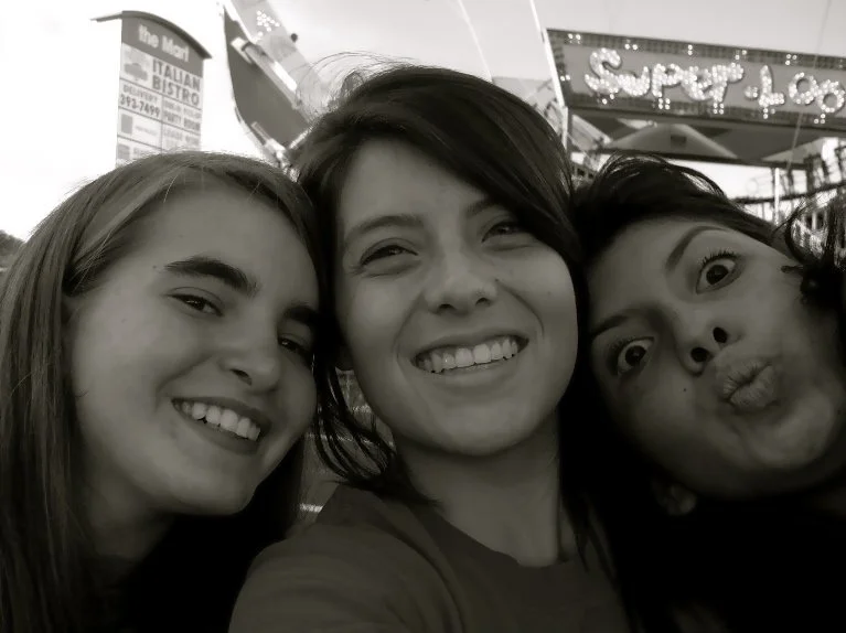 Three women taking a selfie together on a city street, smiling and making silly faces, with signs and lights in the background.