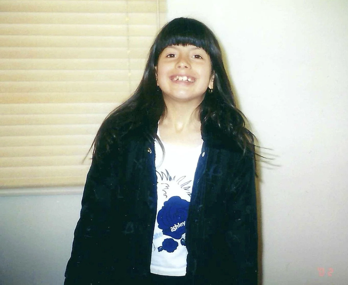 A young girl with long black hair, bangs, and a big smile showing missing front teeth, standing indoors in front of beige blinds. She is wearing a black jacket over a white t-shirt with a dark blue graphic and the name Ashley on it.