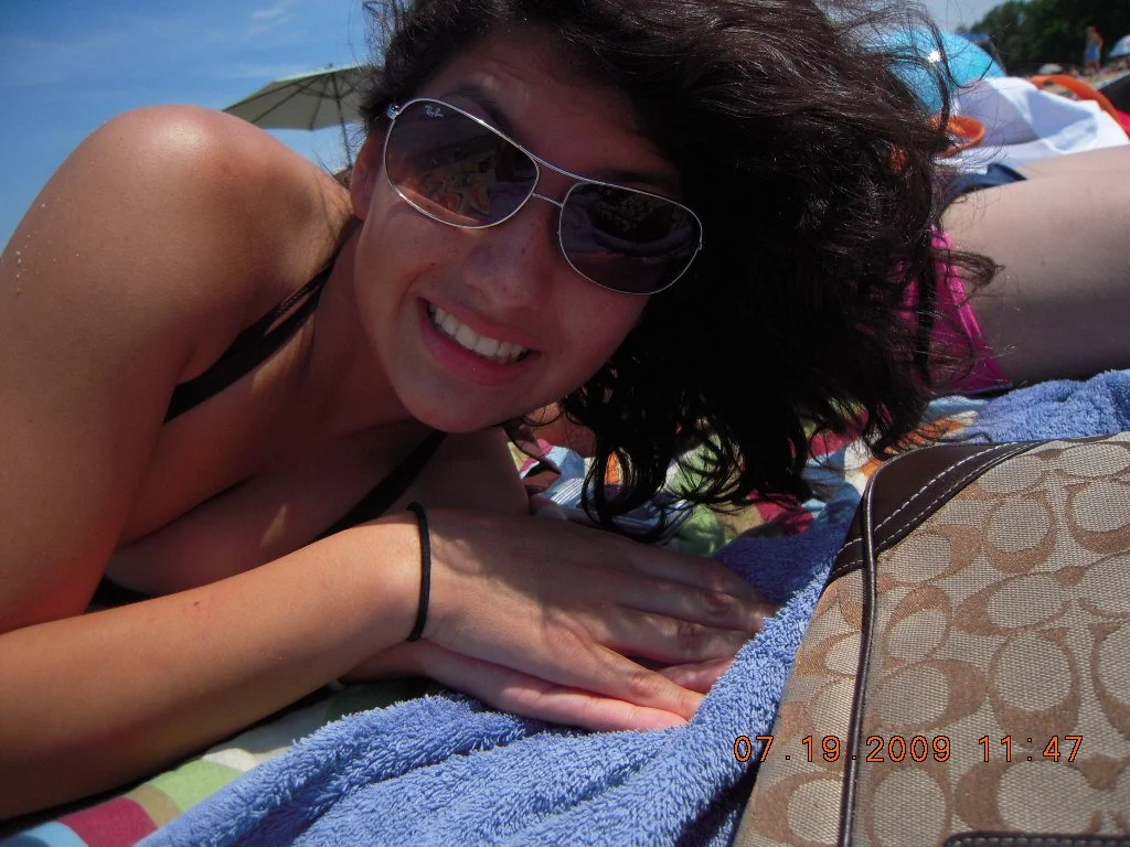 Woman lying on beach towel with sunglasses, smiling, at the beach on a sunny day, with umbrellas and beachgoers in the background.