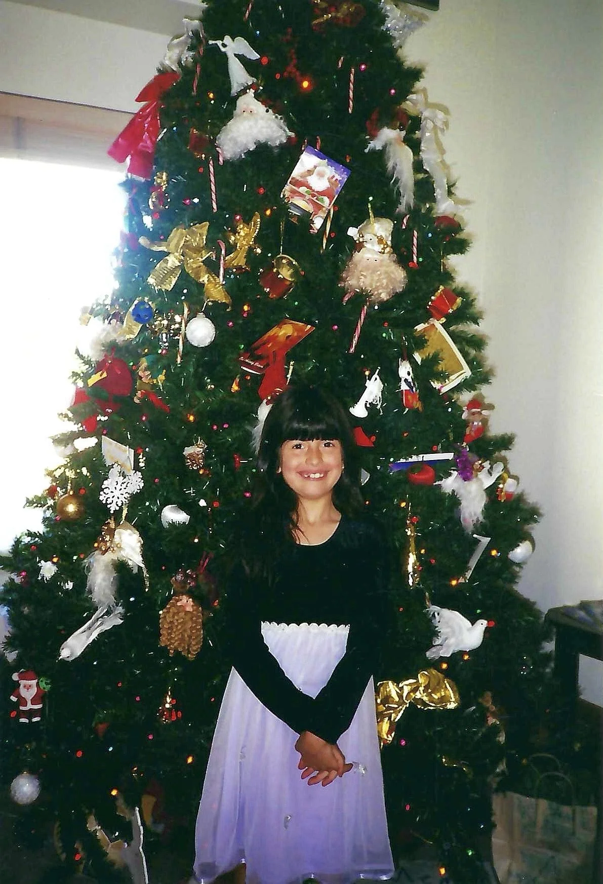 A young girl dressed in a black long-sleeve shirt with a white apron standing in front of a decorated Christmas tree, smiling. The tree is adorned with ornaments, ribbons, and lights.