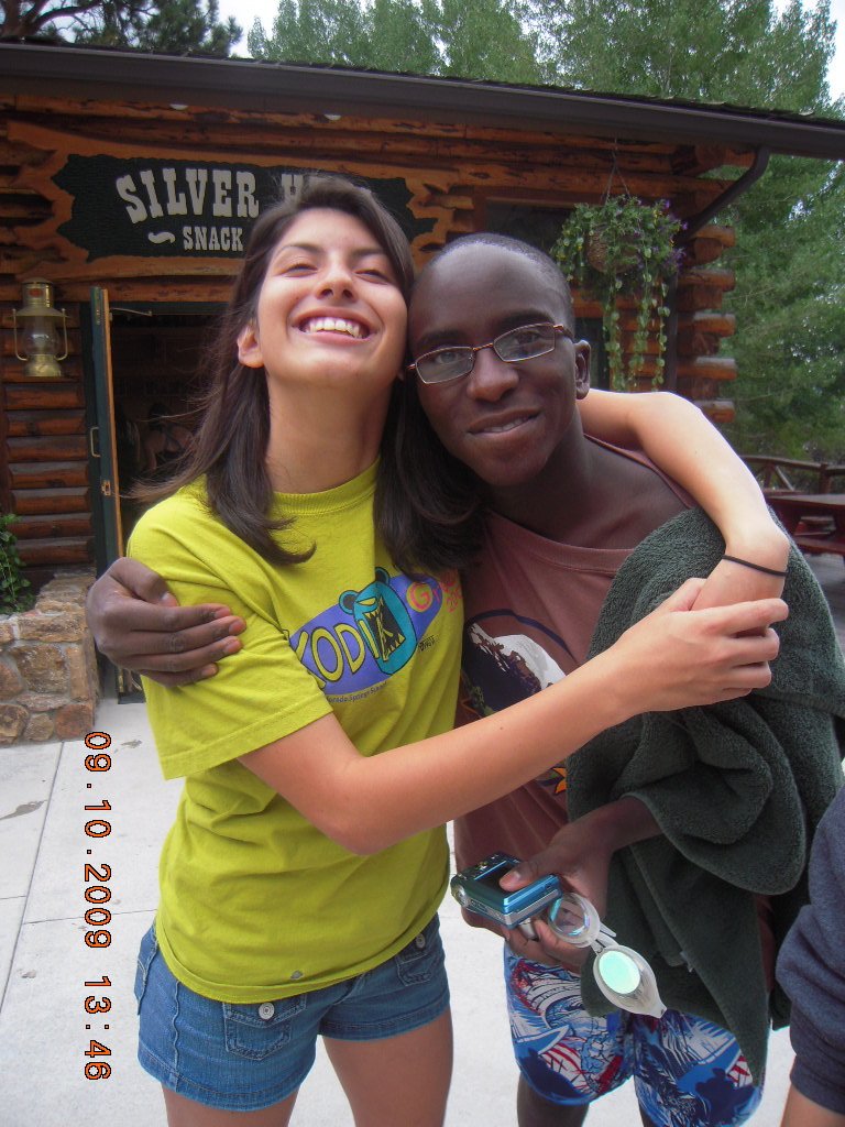 Two young women hugging outdoors, smiling at the camera, one wearing a yellow T-shirt and denim shorts, and the other wearing glasses and colorful shorts, in front of a rustic wooden building with a sign reading 'Silver' in the background.
