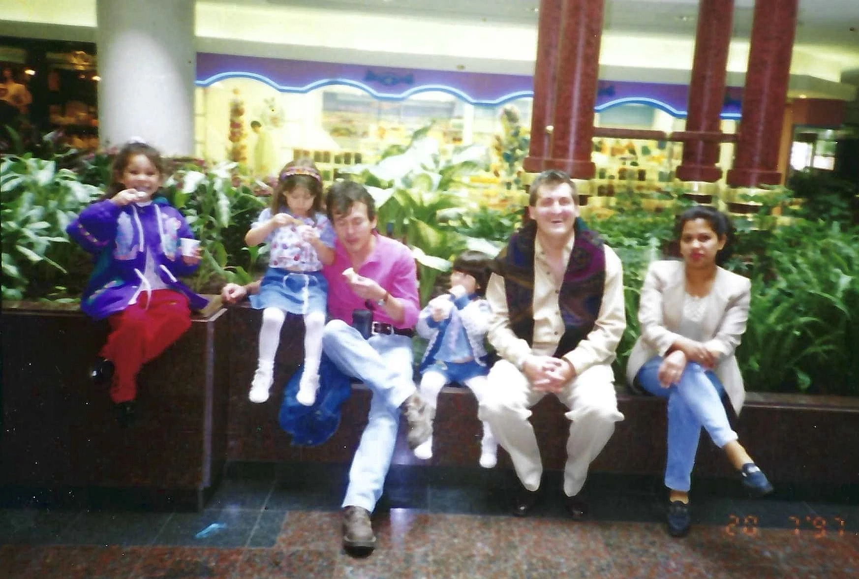 A group of six people, including three children and three adults, sitting on a bench in an indoor setting with plants and decorative columns in the background.