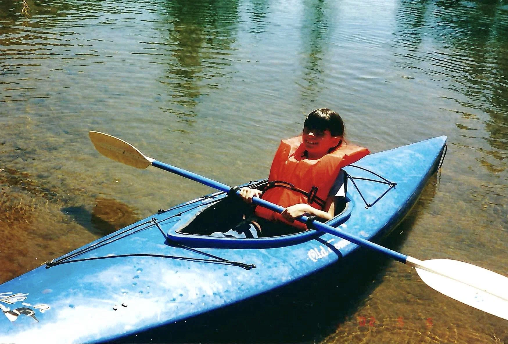 A child in a blue kayak with a red life jacket, paddling on a clear, shallow body of water.