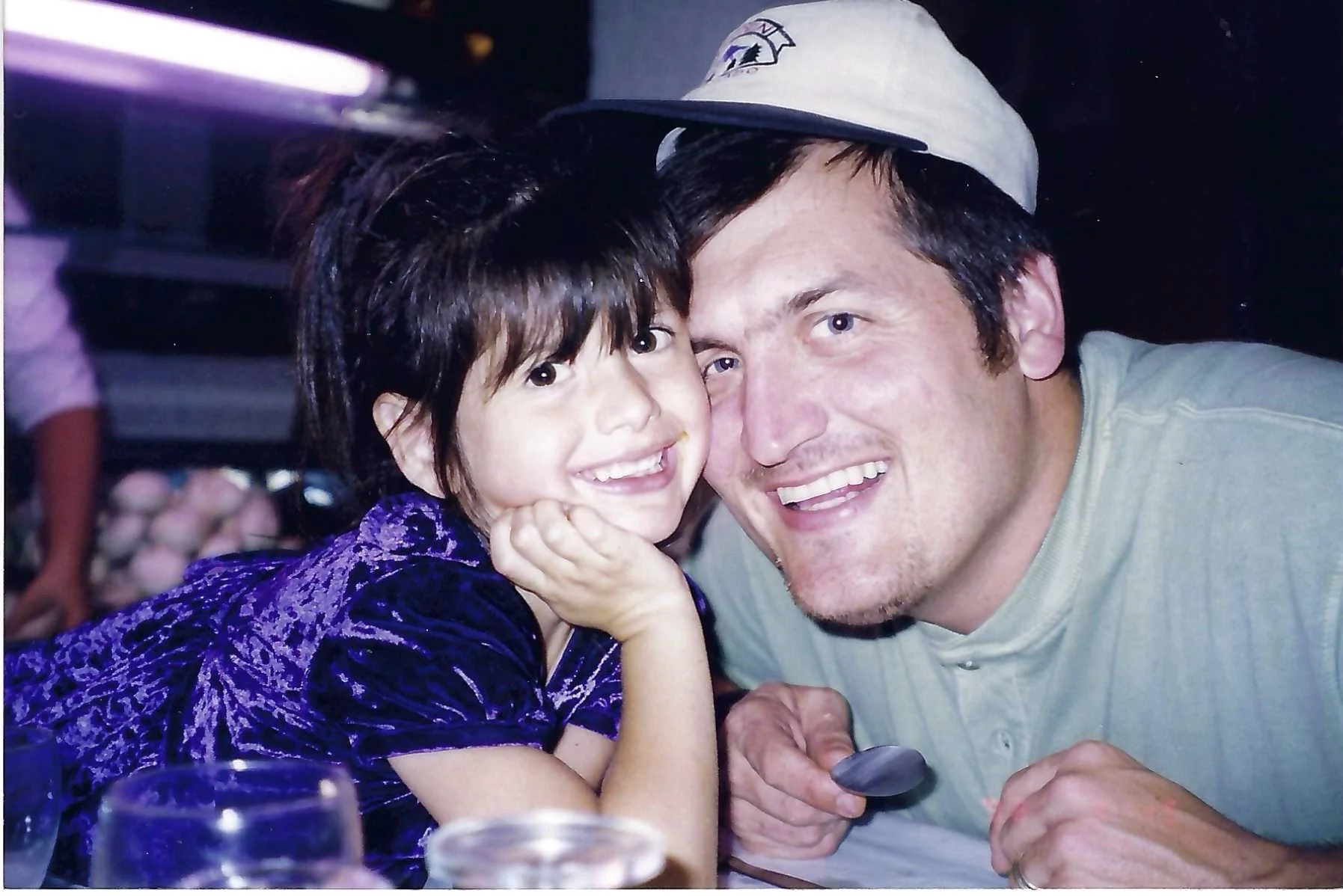 A young girl with dark hair and a purple dress drinks soda while sitting next to a man wearing a beige shirt and a baseball cap, both smiling and close together in a festive setting.
