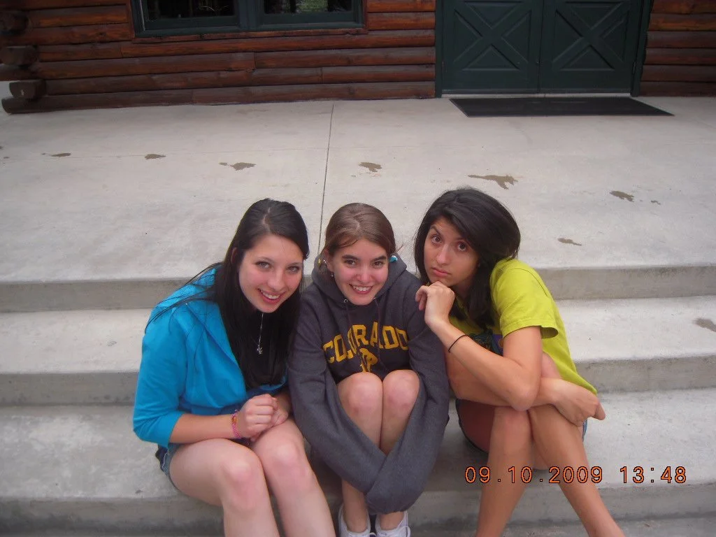 Three young women sitting on concrete steps outside, smiling and looking at the camera.