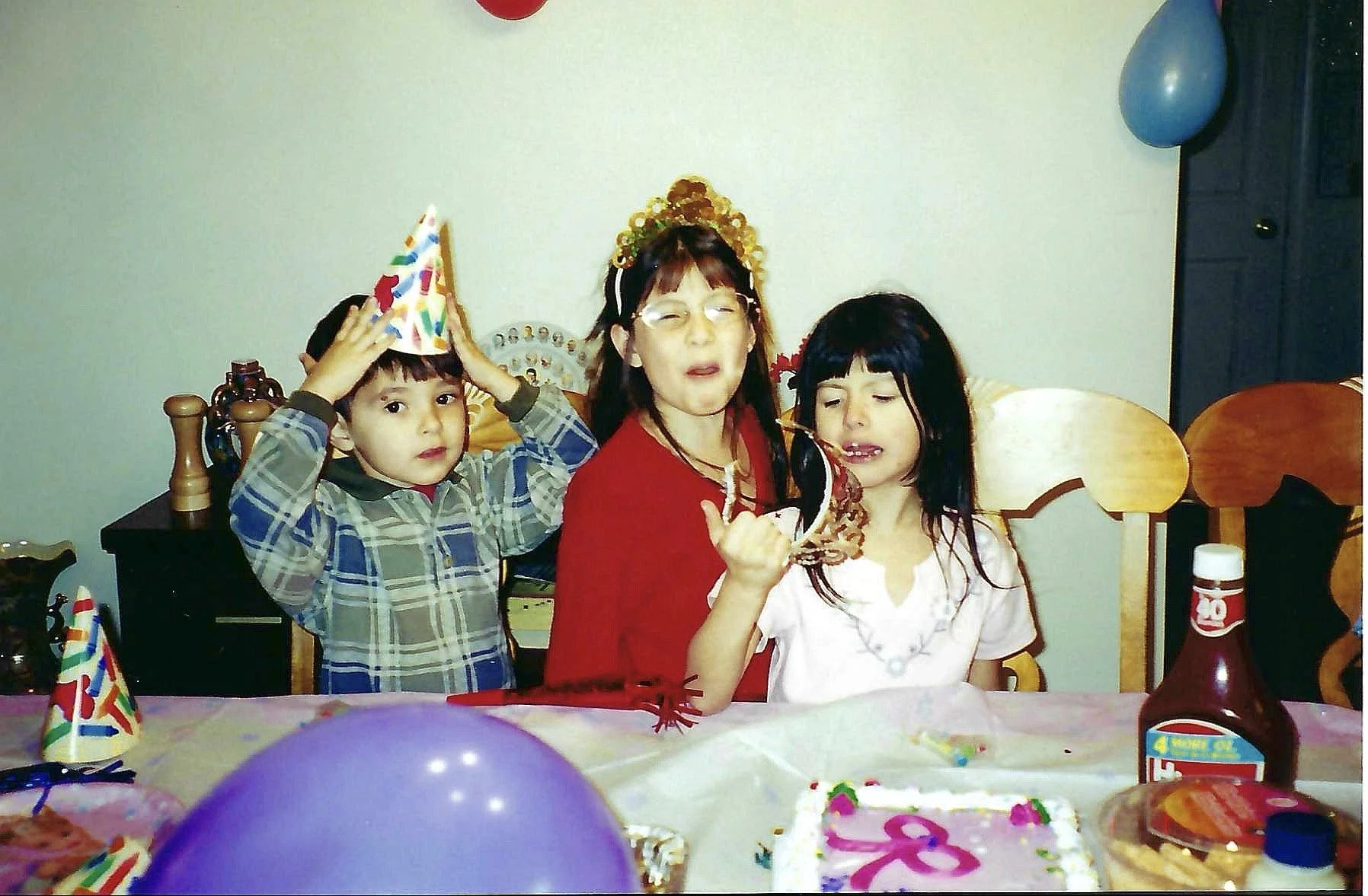 Children and a woman at a birthday party with birthday hats, balloons, and a decorated cake.