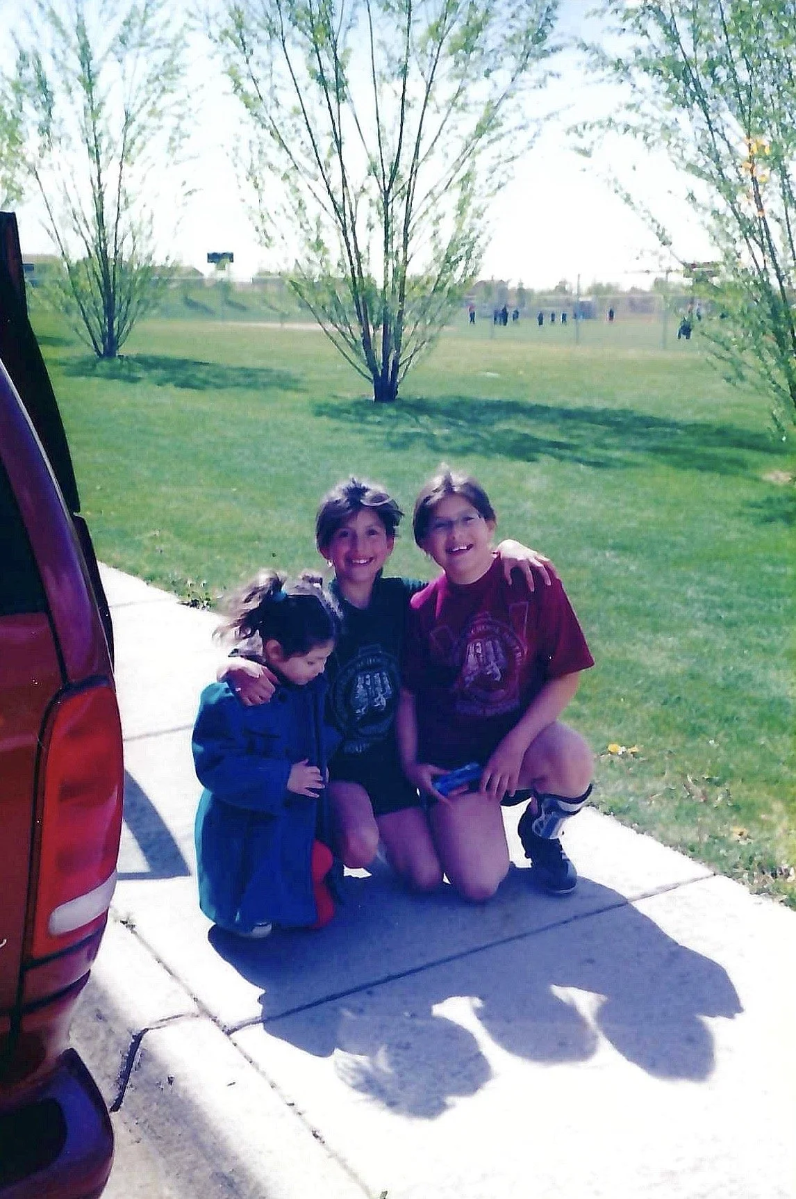 Three children, two girls and one boy, smiling and posing with their arms around each other outdoors on a sunny day, with a grassy field and trees in the background.