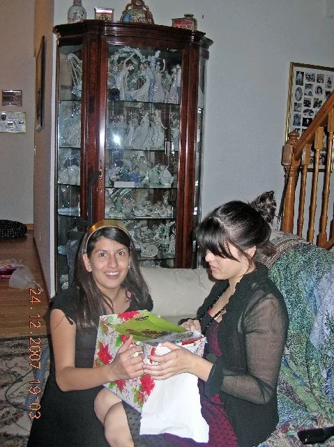 Two women sitting on a couch, one holding a wrapped gift. Behind them is a wooden display cabinet with glass shelves filled with glass figurines and decorative items.