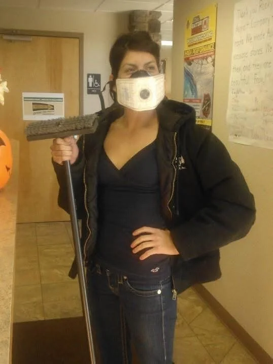 Woman in a black jacket and jeans wearing a face mask holding a mop in a hallway.