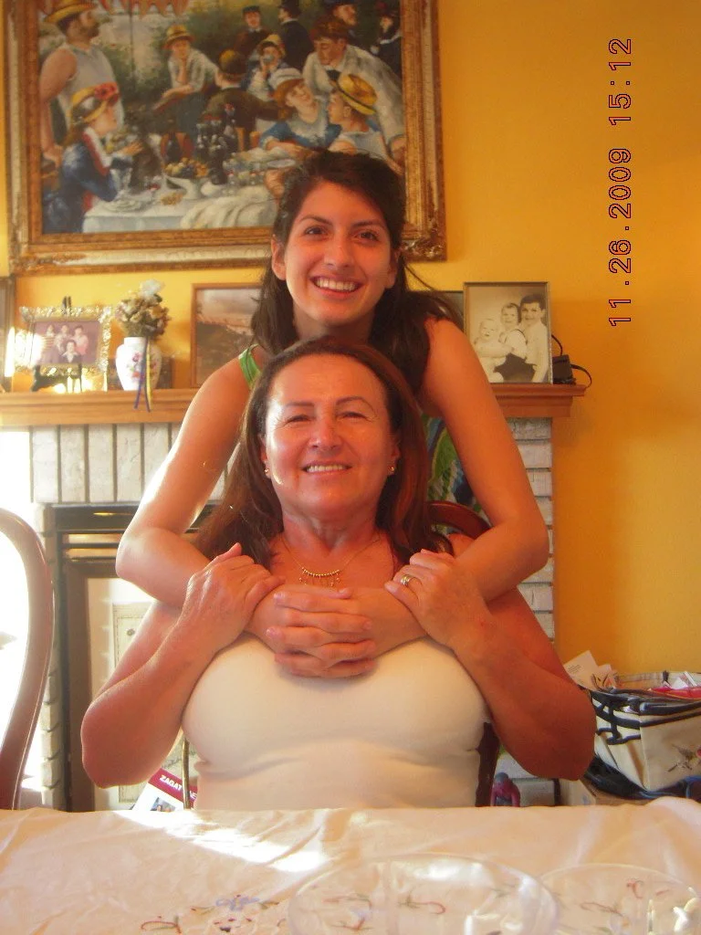 A woman with dark red hair and light skin sitting at a table, smiling and holding hands with a younger woman standing behind her, with dark hair and light skin. They are in a warmly decorated room with a yellow wall, framed photos, and a painting of 