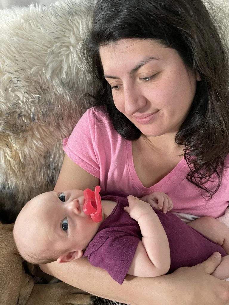 A woman with dark hair wearing a pink shirt holding a baby with a pacifier, lying on her arm on a bed with a fluffy blanket in the background.
