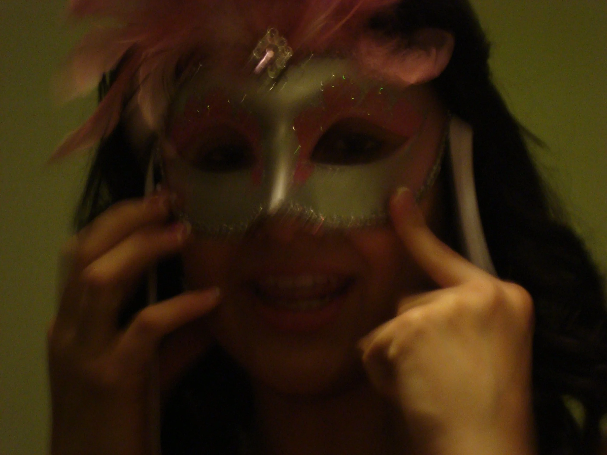 A blurry photo of a person holding a mirror, capturing a reflection of their face painted with clown-style makeup, greenish eyeshadow, red lipstick, and black lipstick, with long dark hair. The background is a plain light-colored wall.