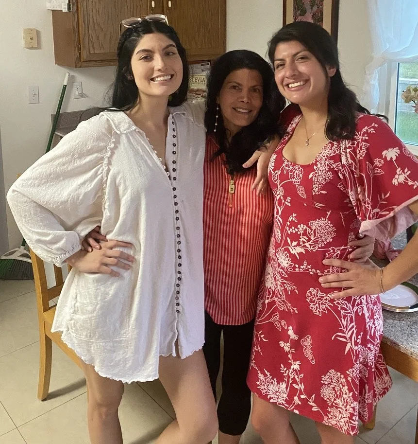 Three women indoors, smiling for the camera, standing close together with arms around each other. One woman on the left wearing a loose white shirt, the middle woman in a red and white striped top, and the woman on the right in a red dress with white