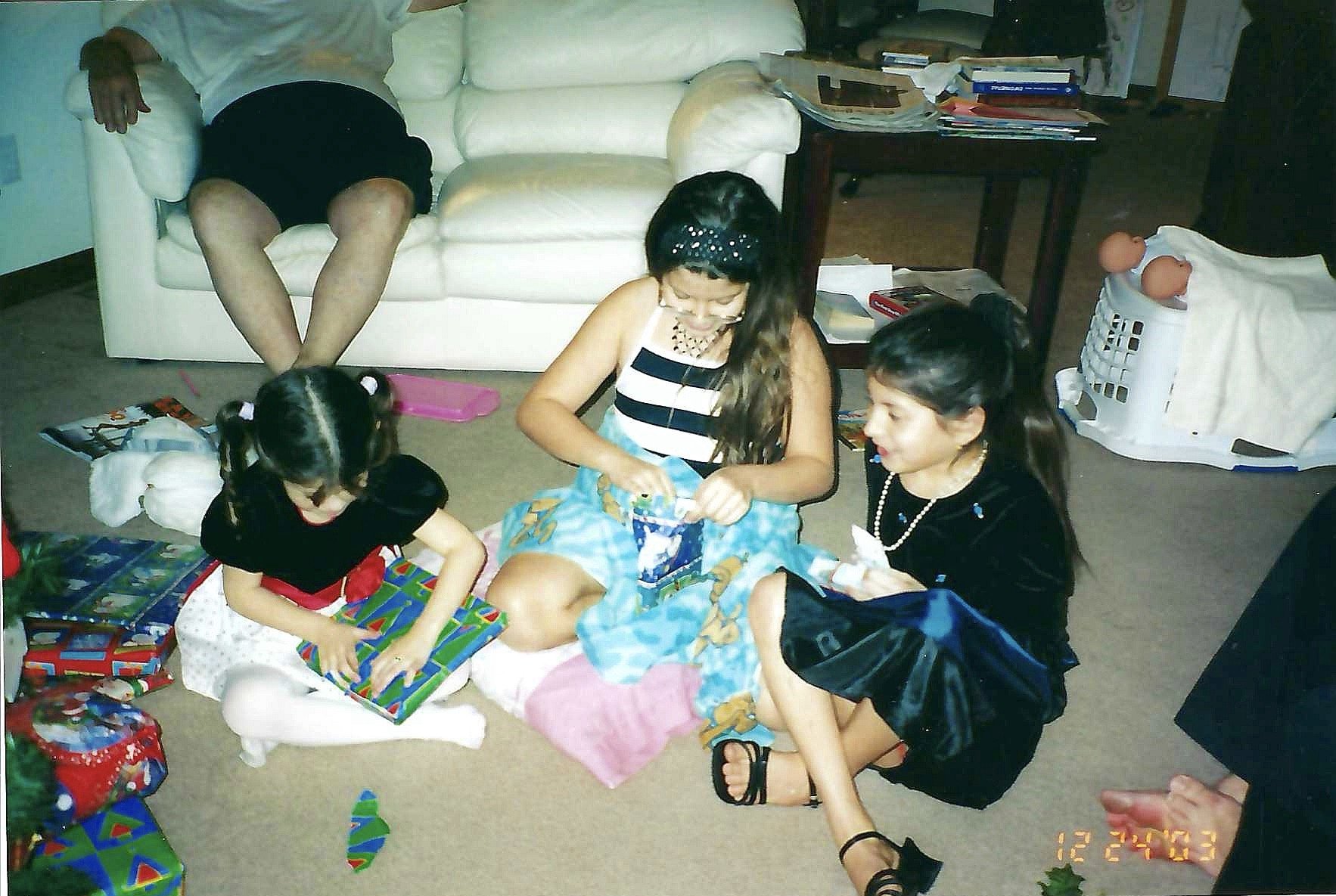 Children opening presents on the floor with an adult sitting nearby, surrounded by gift-wrapped boxes and Christmas decorations in a living room.