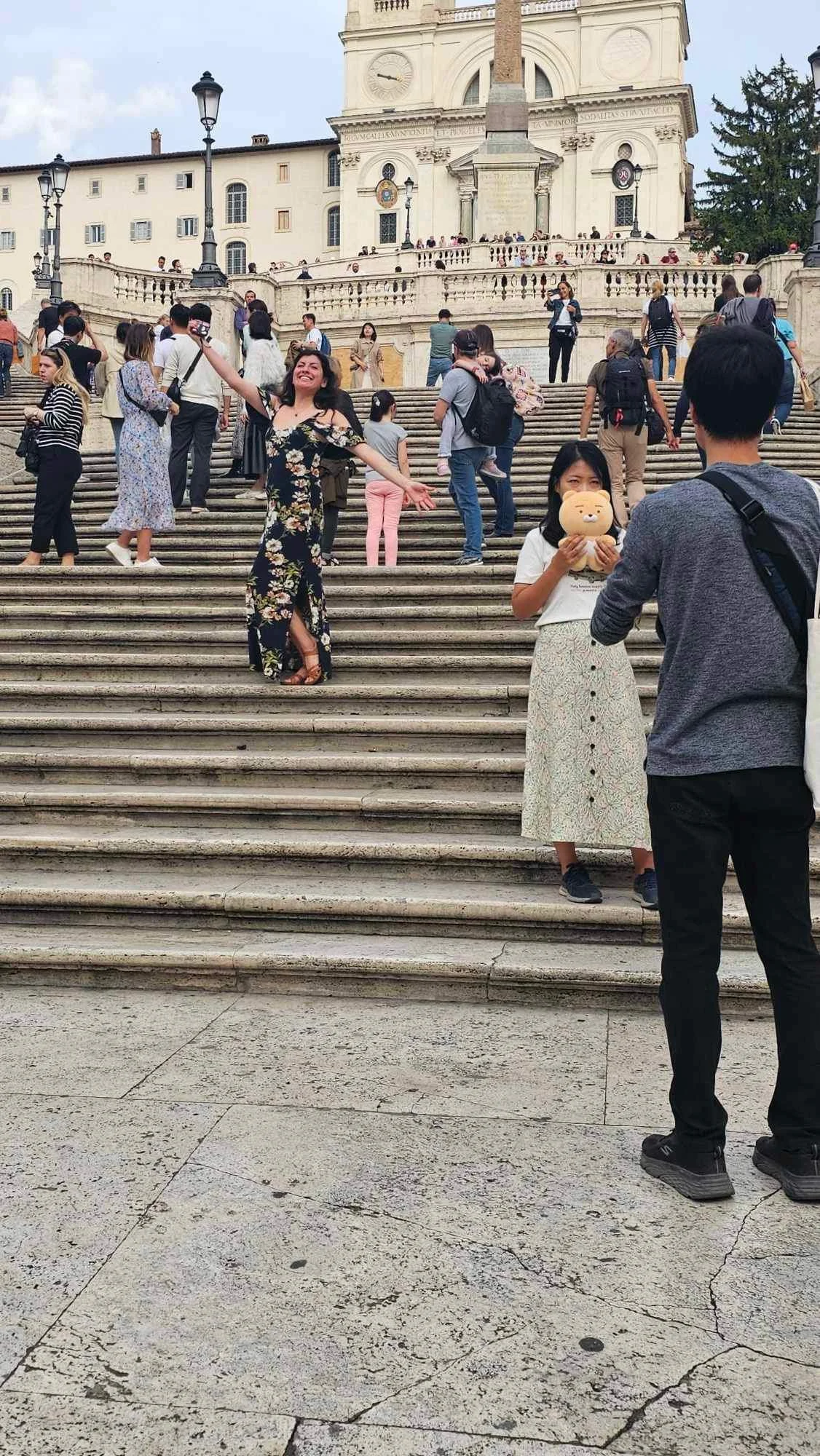 People gathered and taking pictures on the steps of a historical building, with one woman posing and another holding a stuffed animal, in an outdoor setting.