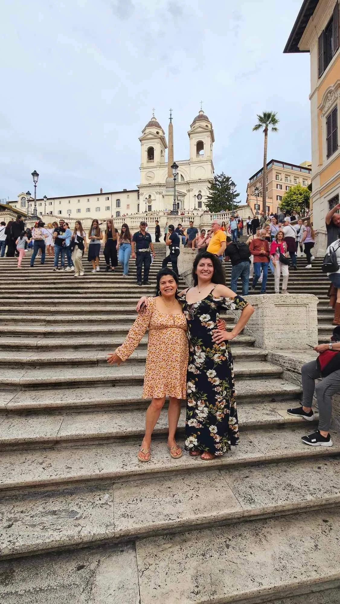 Two women standing on stone steps in front of a crowd at a historic church with twin bell towers in the background, under cloudy sky.