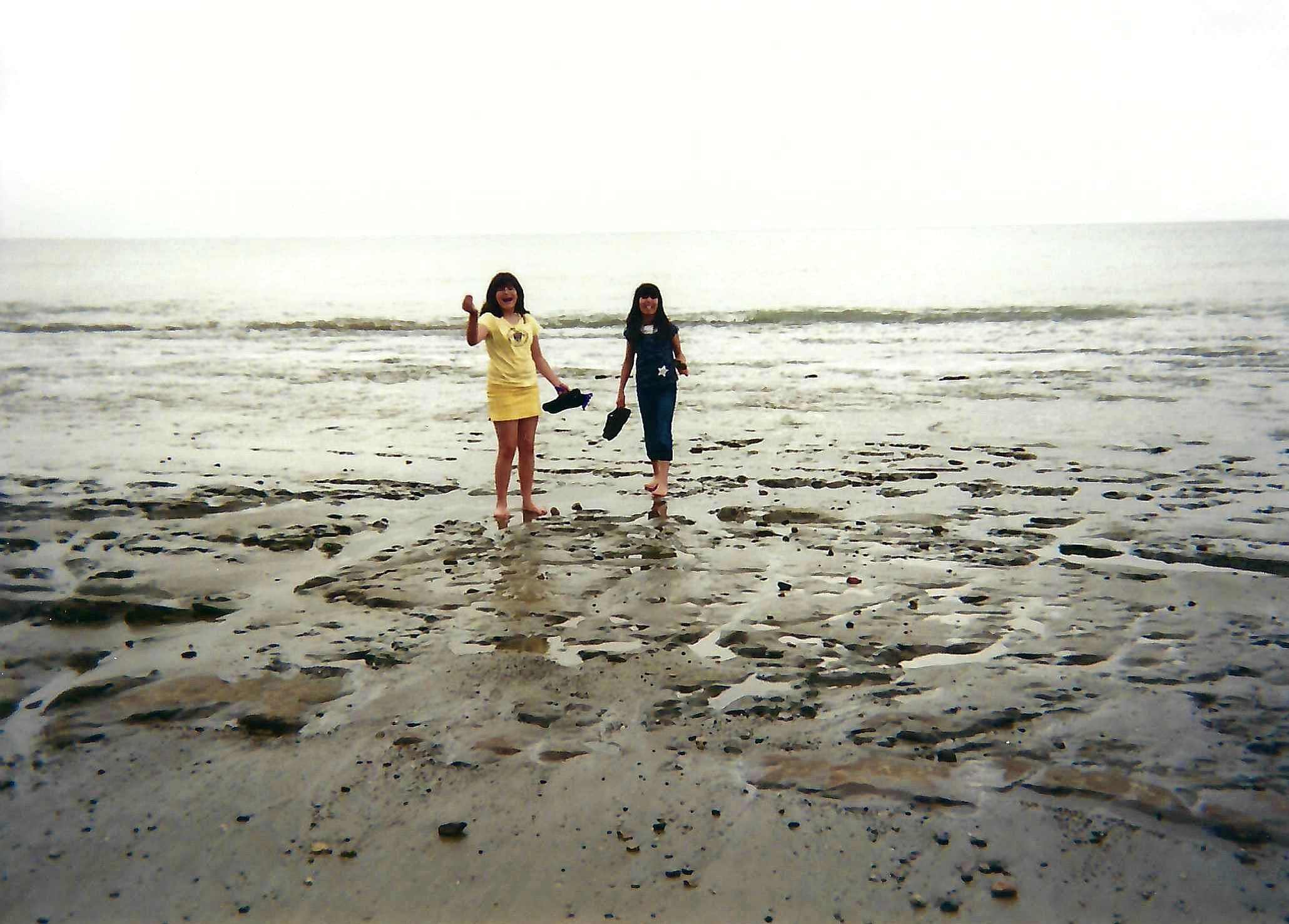 Two young women walking on a sandy beach near the ocean, holding shoes, with waves in the background.
