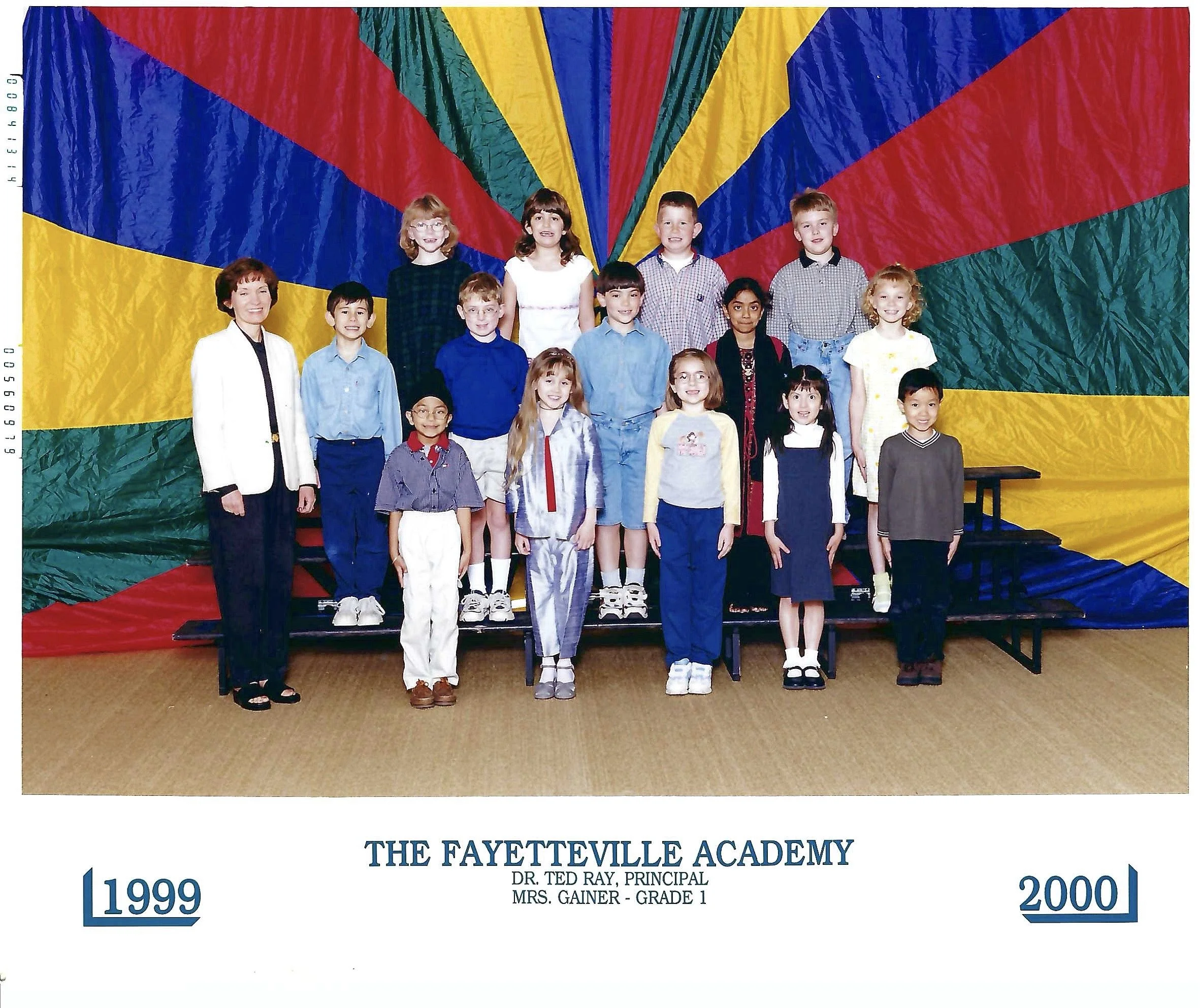 Group of diverse children and a teacher standing in front of a colorful fabric backdrop at Fayetteville Academy graduation ceremony, with years 1999 and 2000 displayed at the bottom, indicating a graduation event.