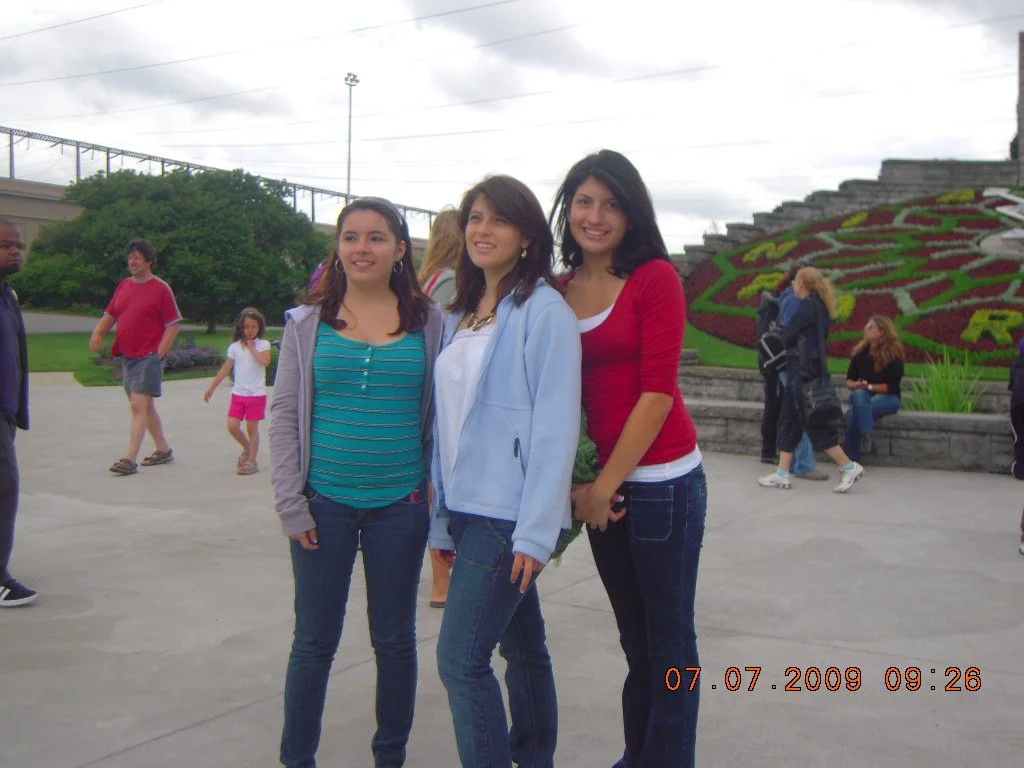 Three young women standing together outdoors on a cloudy day, with a flower bed and other people in the background.