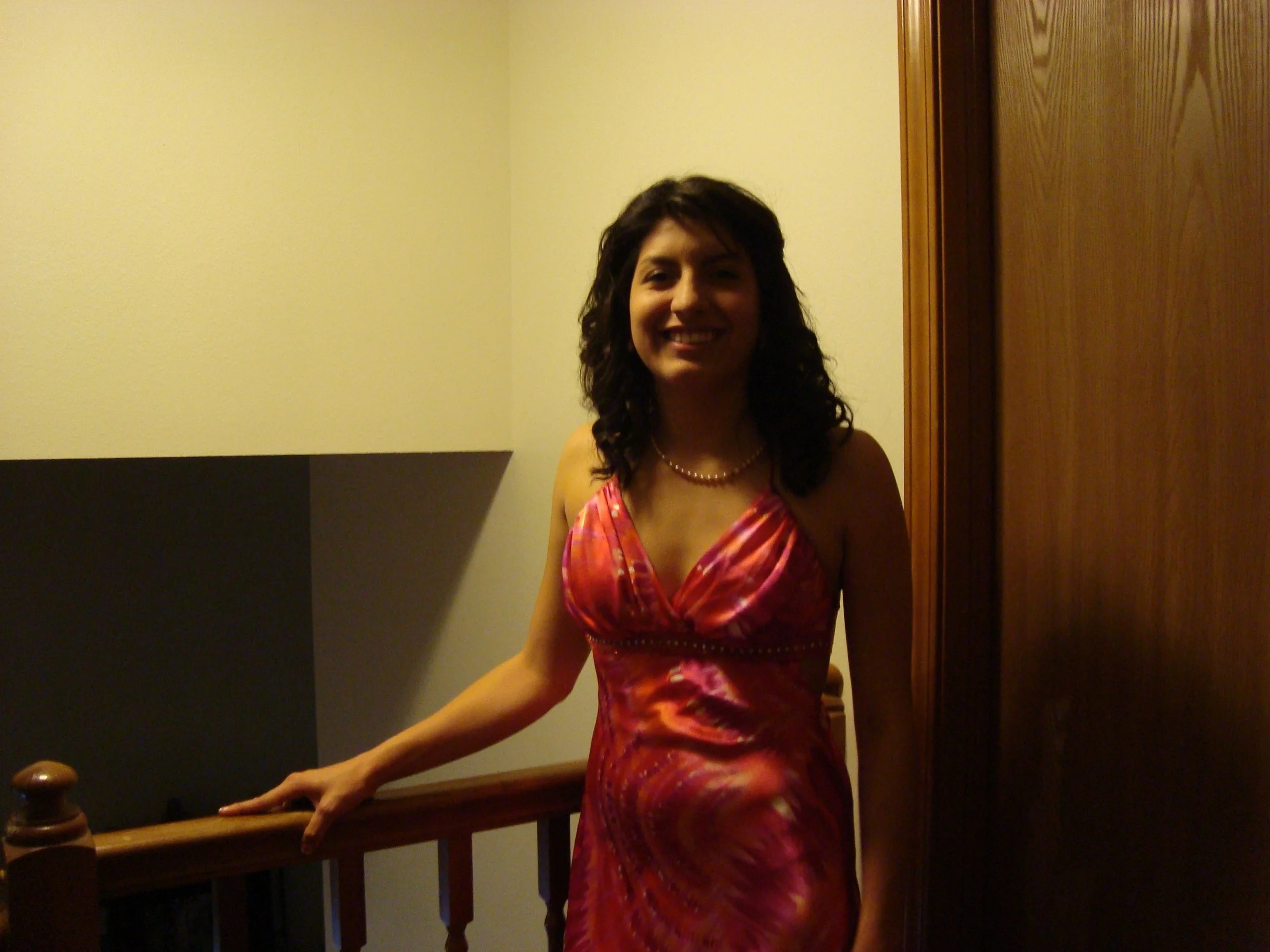 A woman wearing a pink and purple dress with a pearl necklace, smiling, standing indoors next to a wooden panel and a beige wall.