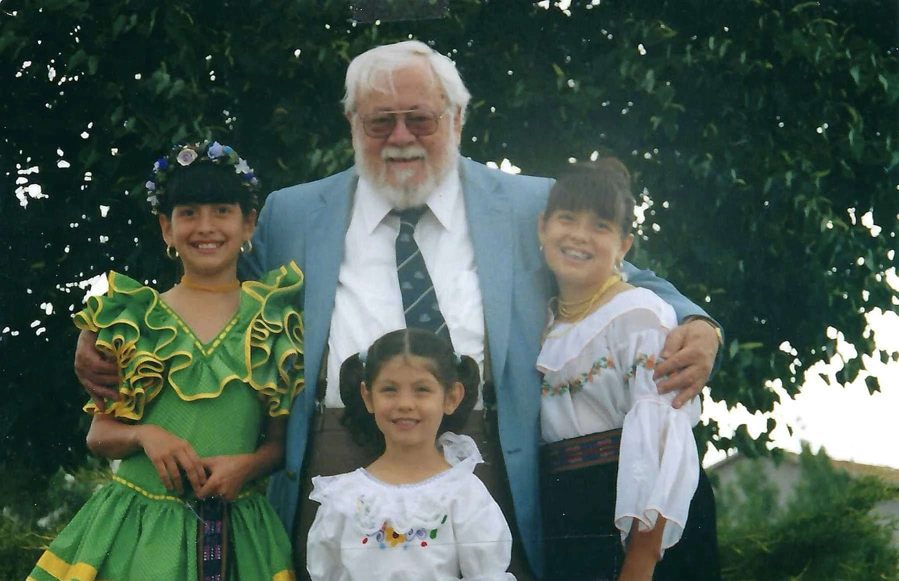 A group of five people enjoying a day outdoors with a large tree in the background, including a man with white hair and beard in a blue blazer, and four young girls dressed in colorful traditional Mexican attire.