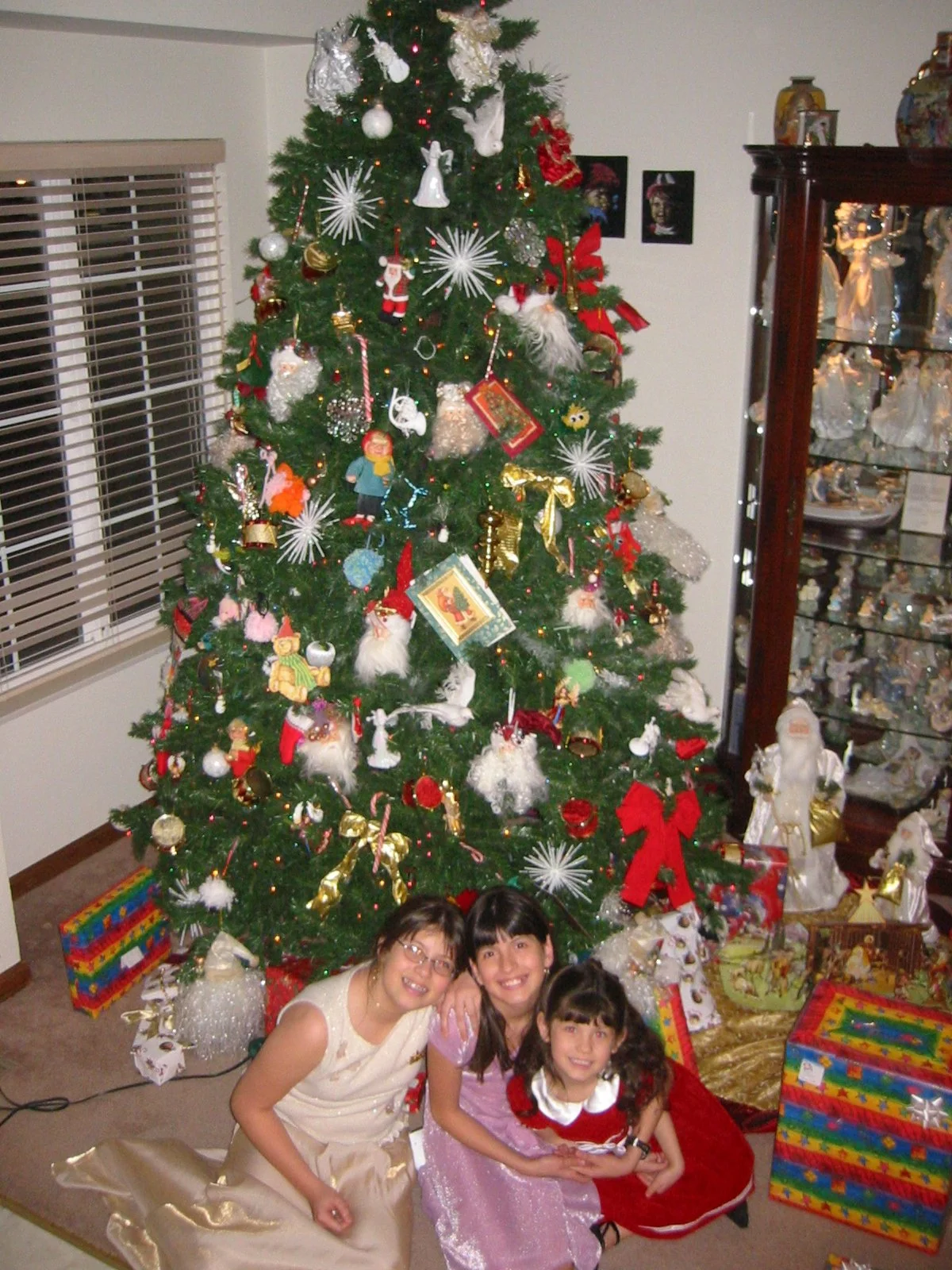A decorated Christmas tree with ornaments, ribbons, and presents underneath. Three women and a girl are sitting on the floor in front of the tree, smiling. There's a window with blinds to the left and a glass cabinet filled with Christmas decorations