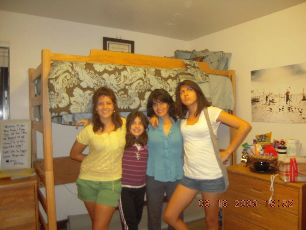 Four young women standing together in a bedroom, smiling at the camera, with a bed and a dresser in the background.