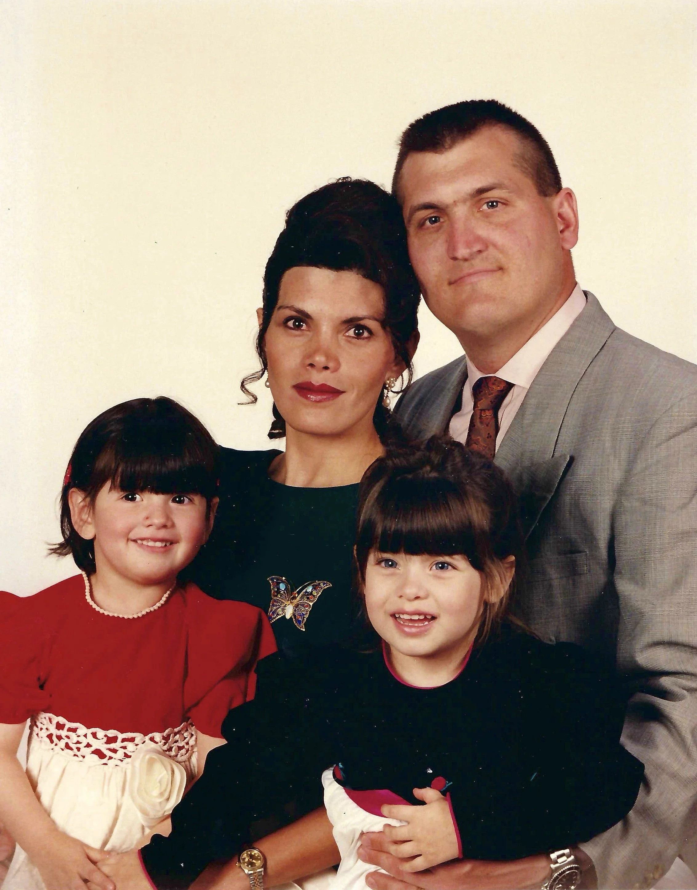 A family group portrait with two adults and three children, all dressed in festive attire, smiling and standing against a plain background.