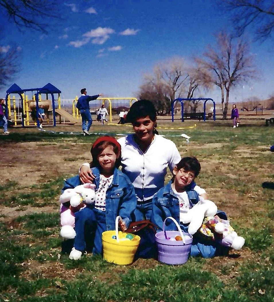 A woman and two children with Easter baskets in a park during spring, with a playground and trees in the background, on a sunny day.