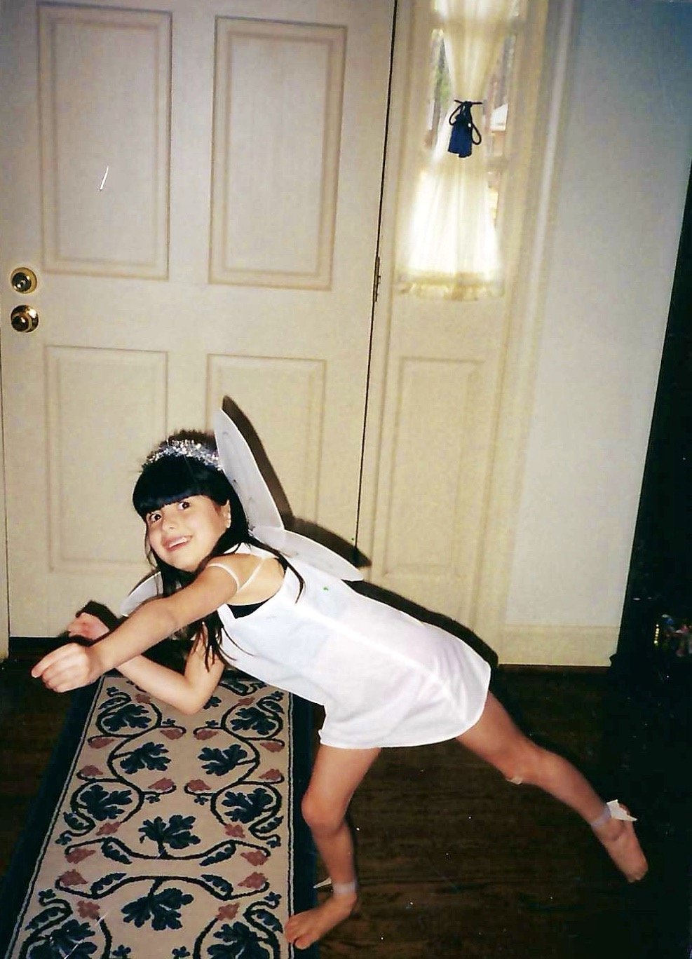 A young girl dressed as a fairy, wearing a white dress, fairy wings, and a tinsel headband, posing playfully in front of a door inside a home.