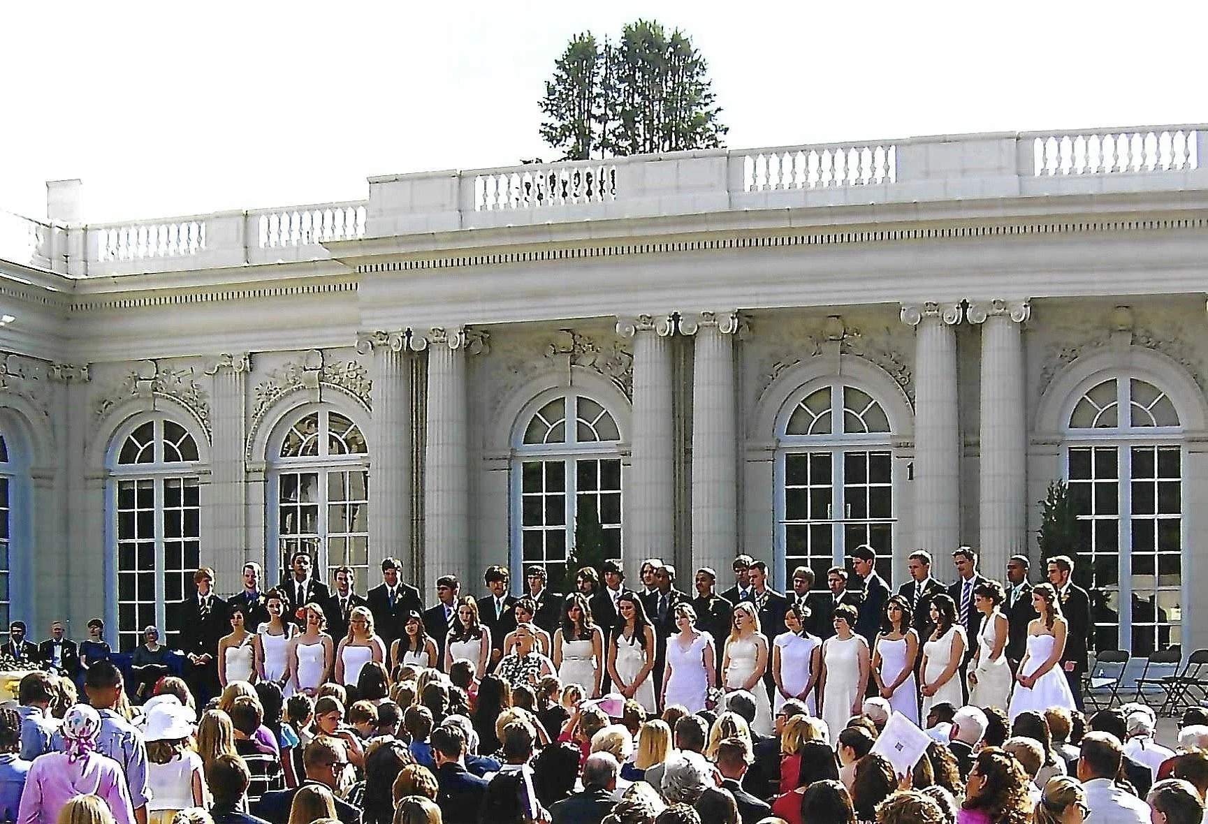 A large group of graduates dressed in formal attire, standing in front of a grand, ornate building with large arched windows, taking part in a graduation ceremony.