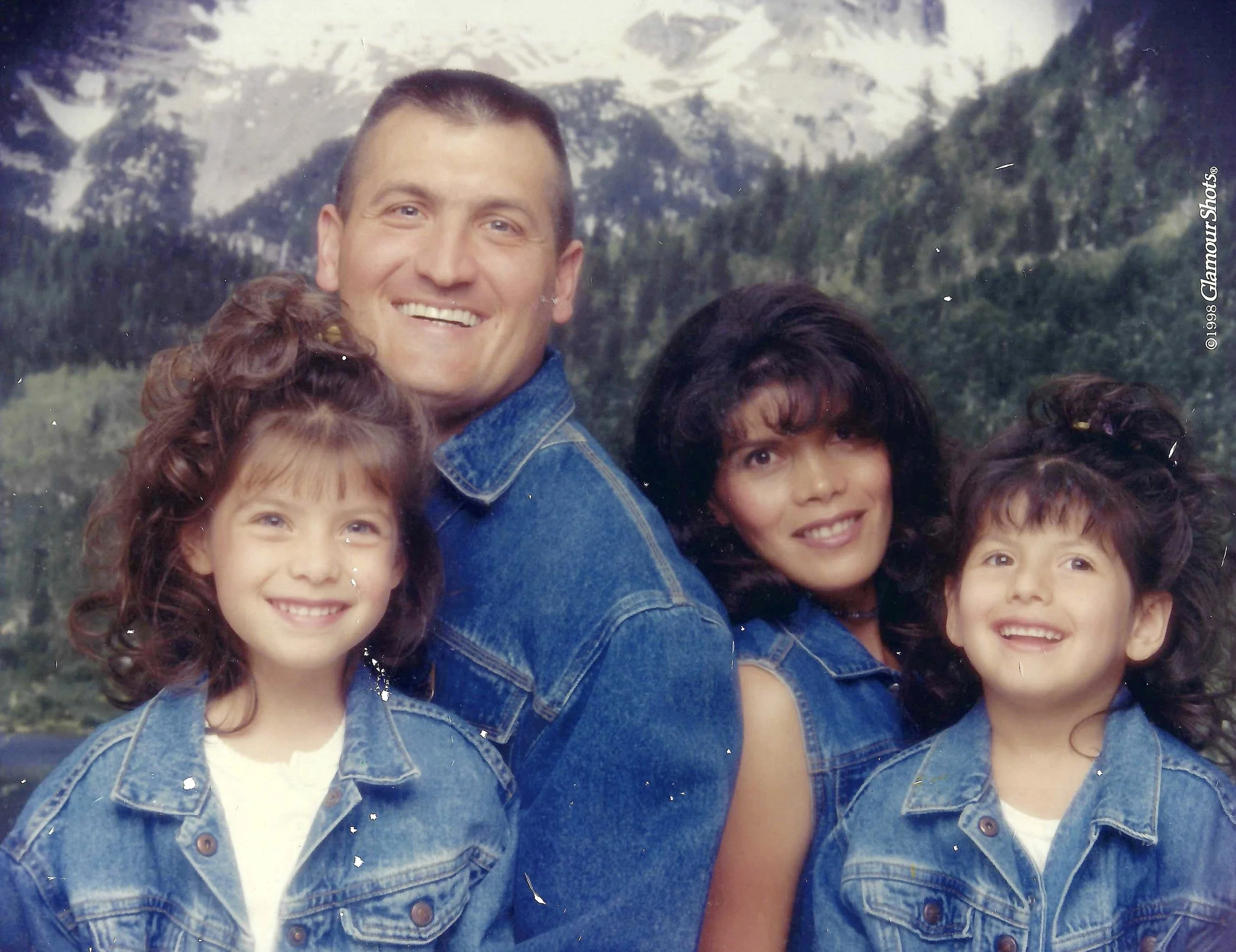 A group of smiling people wearing denim jackets outdoors in front of a mountain landscape.