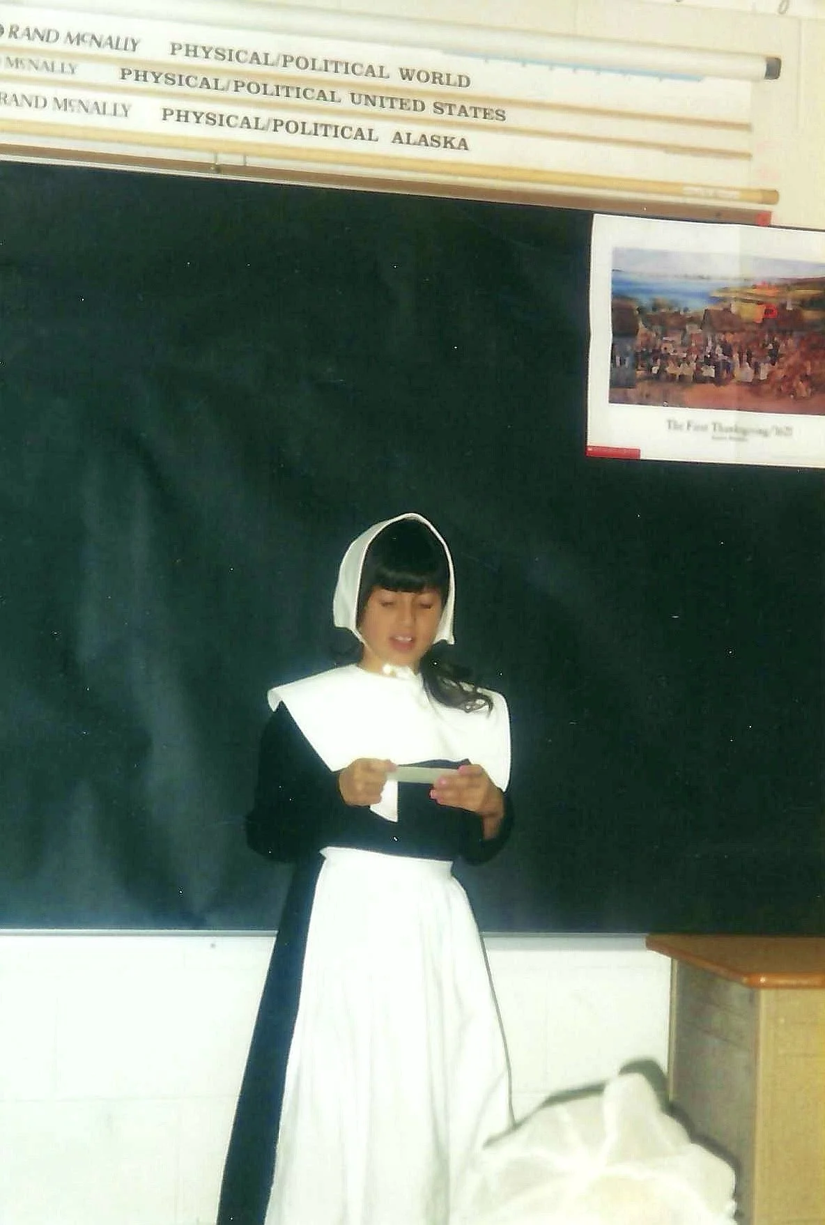 A woman dressed as a nurse in a black and white costume standing in front of a blackboard in a classroom. She is holding a paper, with books on political and world history on a shelf above her and a poster on the wall.