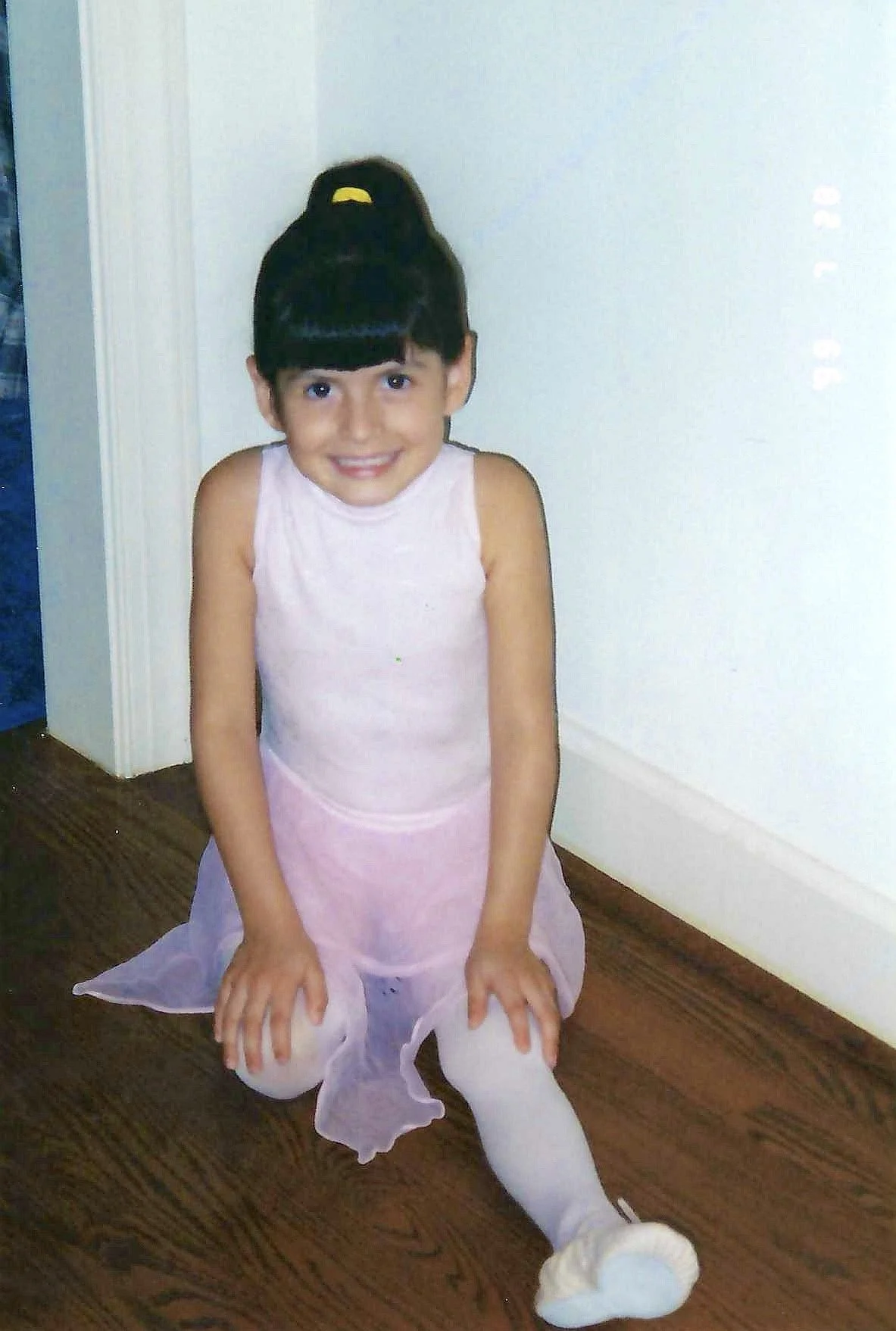 A young girl with a bun hairstyle decorated with a yellow bow, dressed in a pink ballet outfit, sitting on the floor with legs stretched out, smiling at the camera in a room with wooden flooring and light-colored walls.