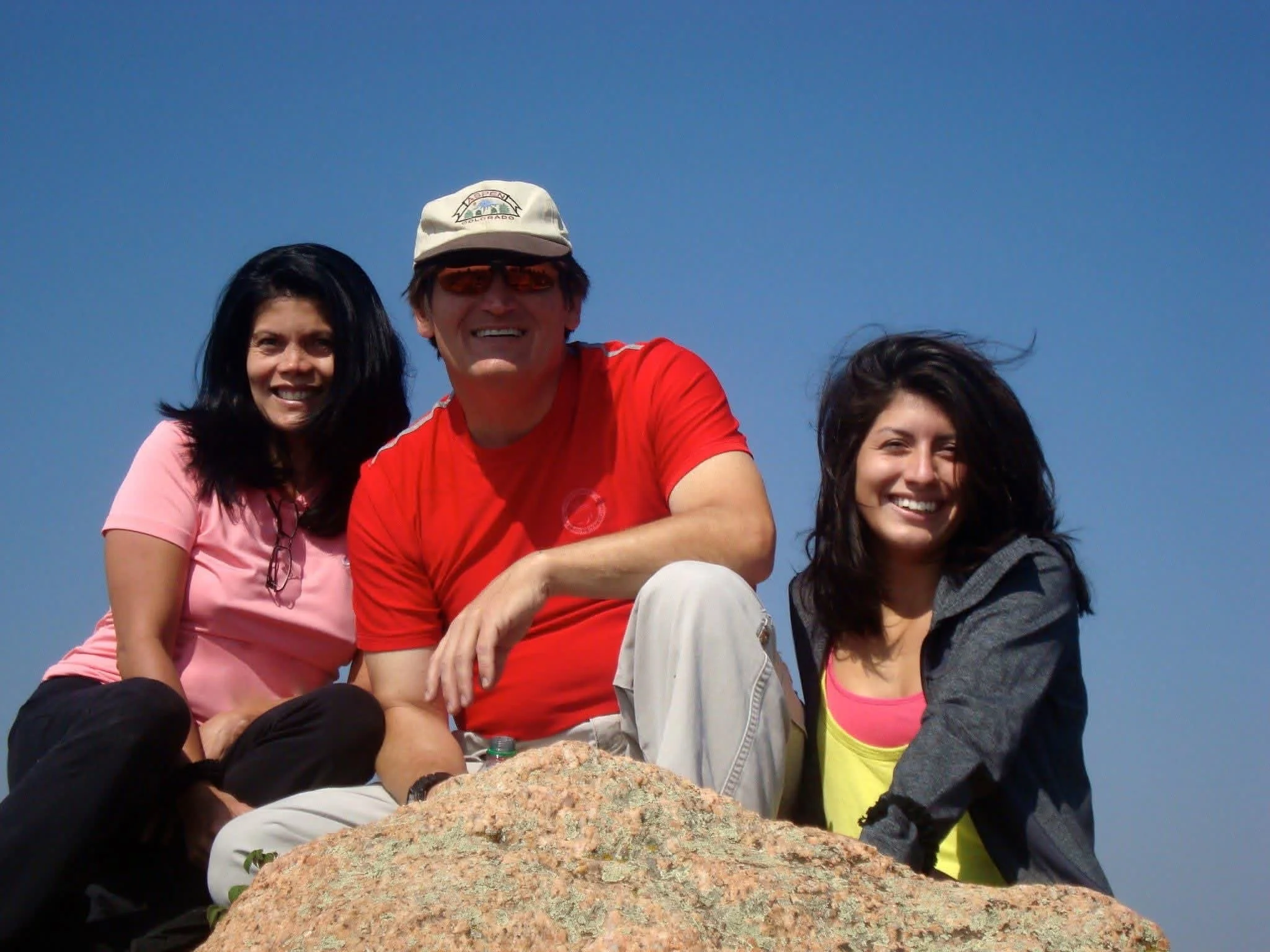 Three smiling people sitting on a large rock outdoors against a clear blue sky, dressed casually, with a man in a red shirt and a woman in a pink shirt in the center.