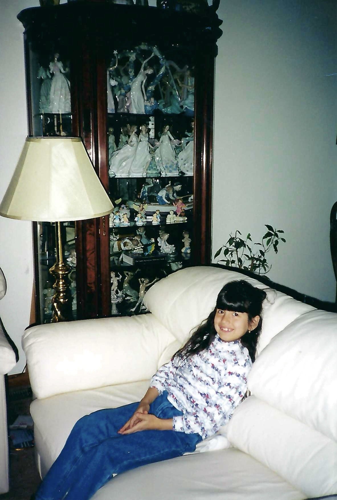 A young girl with dark hair and wearing a floral top, smiling and sitting on a white couch in a living room. Behind her is a tall display cabinet filled with porcelain figurines and decorative collectibles, a lamp with a beige shade, and a small pott