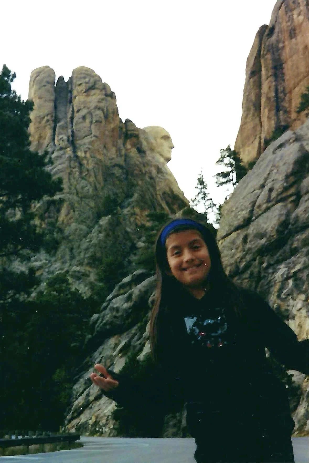 A person with long hair wearing a blue headband and black clothing standing in front of the Mount Rushmore sculpture, with trees and rocky cliffs surrounding.