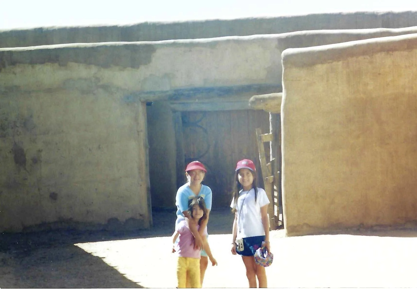 Three children standing outside a rustic clay or adobe building with an open doorway. Two girls are wearing pink hats, and one girl is holding a small dog.