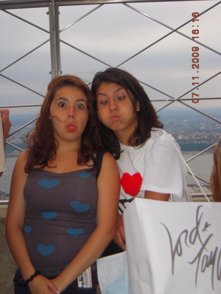 Two young women with surprised and pouting facial expressions, standing in front of a metal observation tower with a cityscape and water in the background.