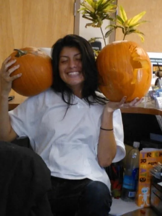 A woman with long dark hair smiling while holding two carved pumpkins on her shoulders in an indoor setting.