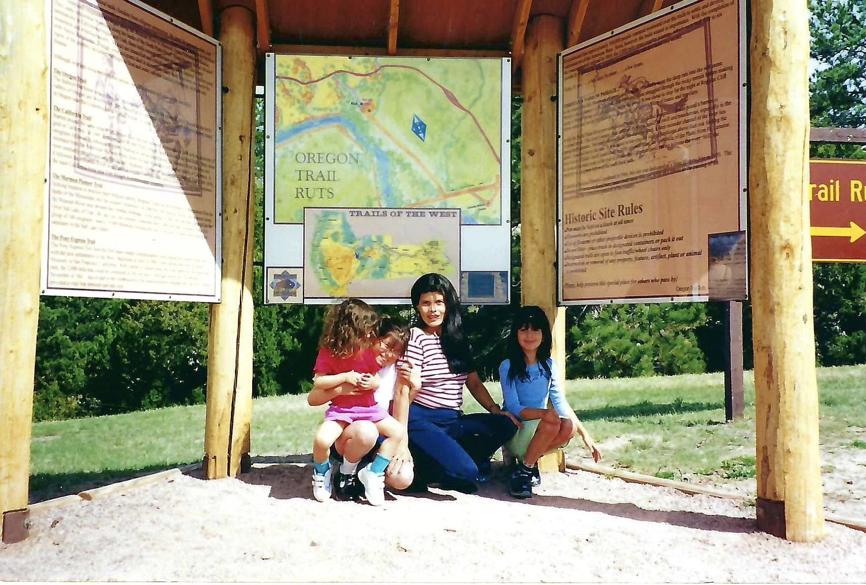 A woman with three young girls sitting on a bench under a wooden shelter at a park, with informational signs and maps about Oregon Trail routes behind them.