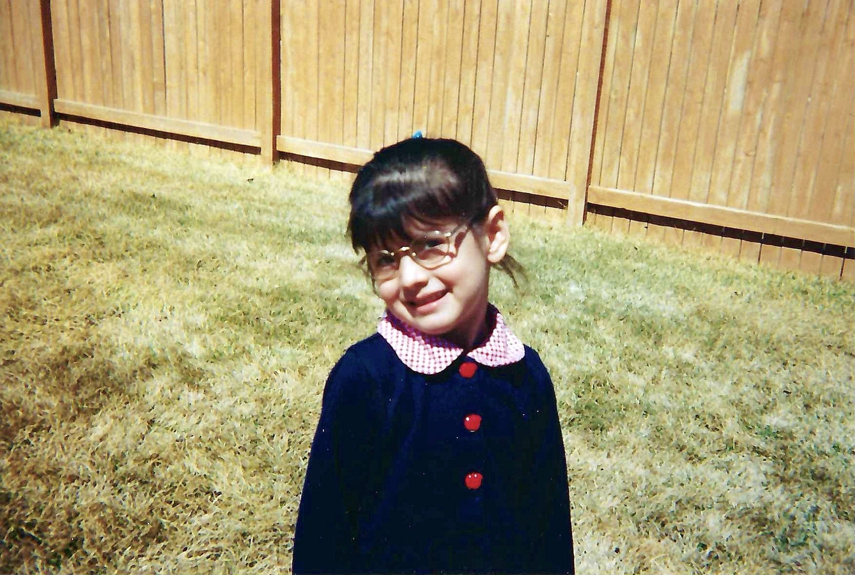 A young girl with glasses standing on a grassy backyard with a wooden fence in the background, wearing a navy blue dress with red buttons and a pink checkered collar.