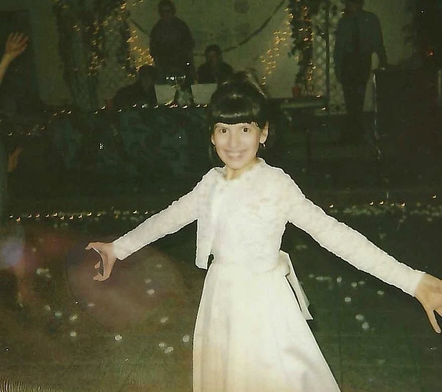 Young girl in a white dress smiling with arms outstretched at a party or event with dim lighting and people in the background.